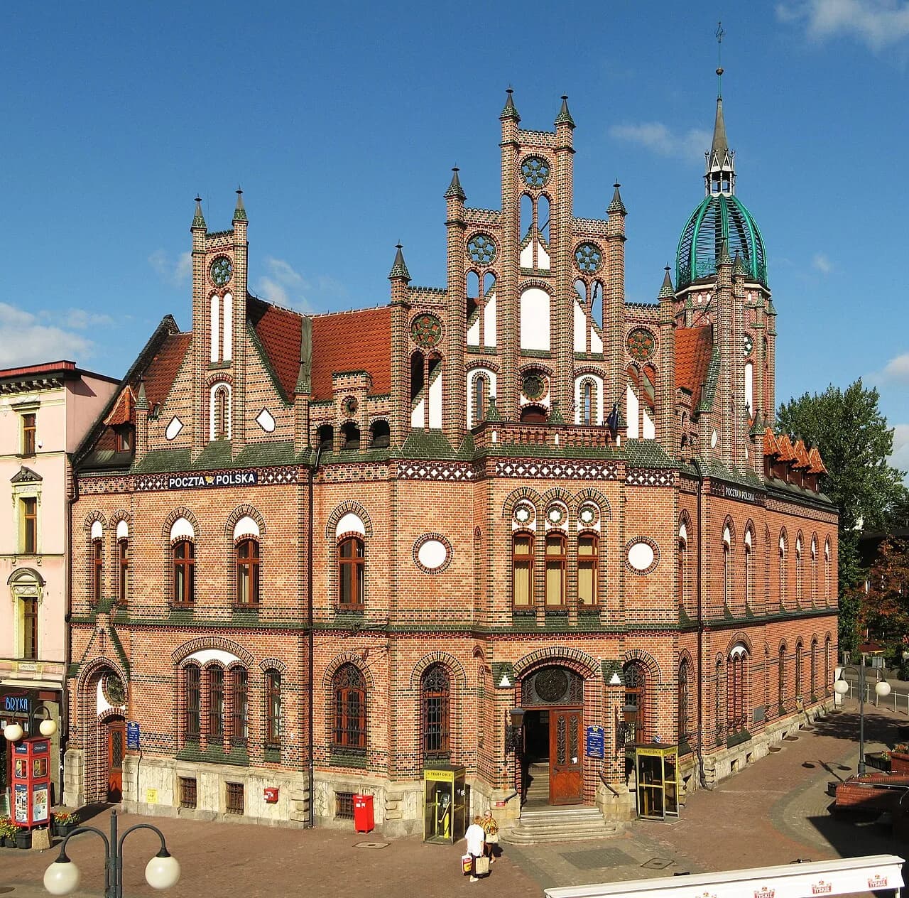 Ornate red brick building with many windows, decorative arches, and a green domed tower with a spire against a clear blue sky, with a red mailbox and streetlights in the foreground.