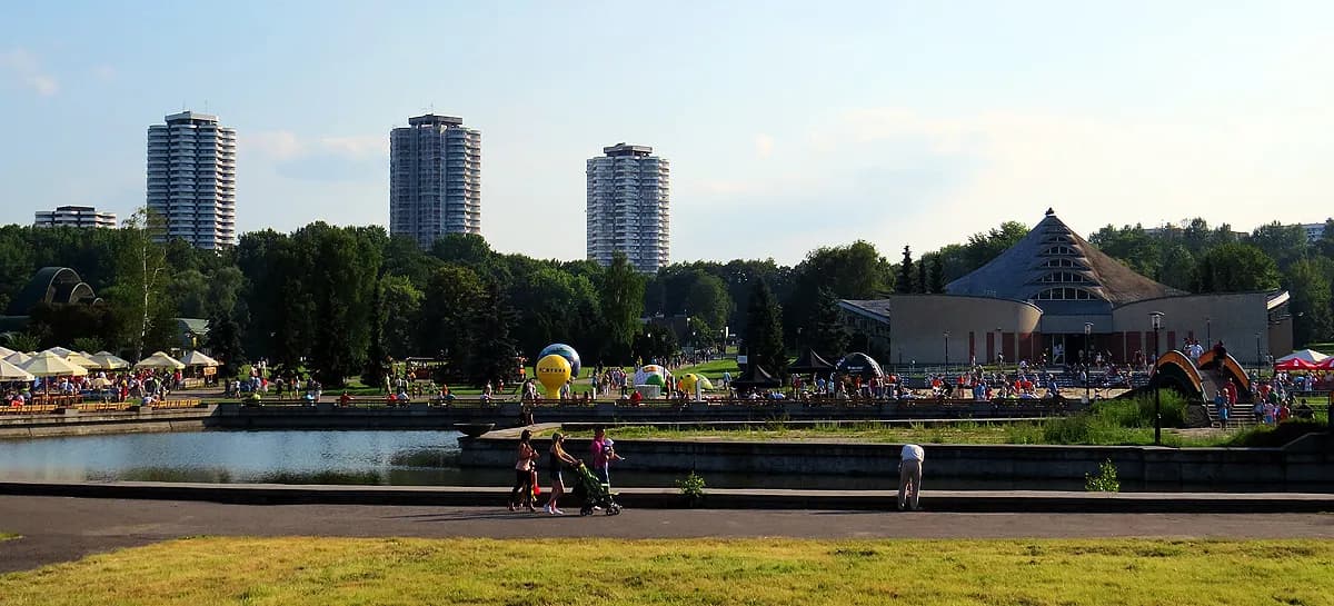 A wide shot of a public park with a pond, several tall buildings in the background, and a large, pyramid-shaped building on the right, all surrounded by trees and people enjoying the sunny weather.