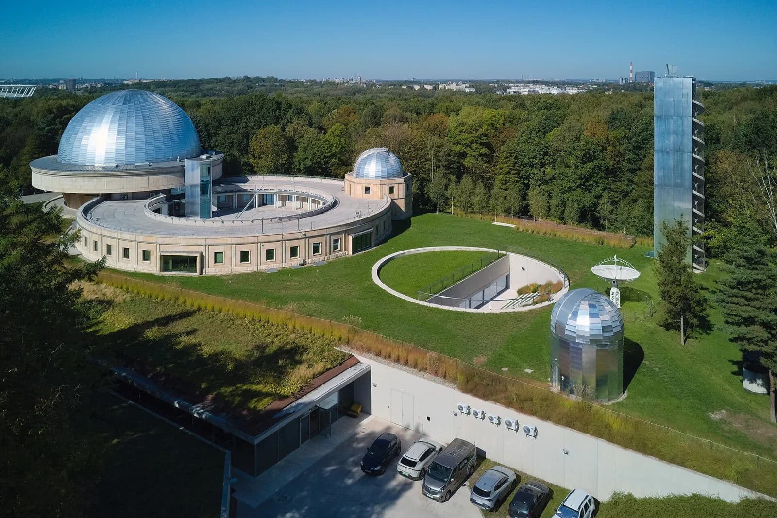 An aerial view of an observatory with a large, silver domed building and a circular complex surrounded by green lawns and trees.