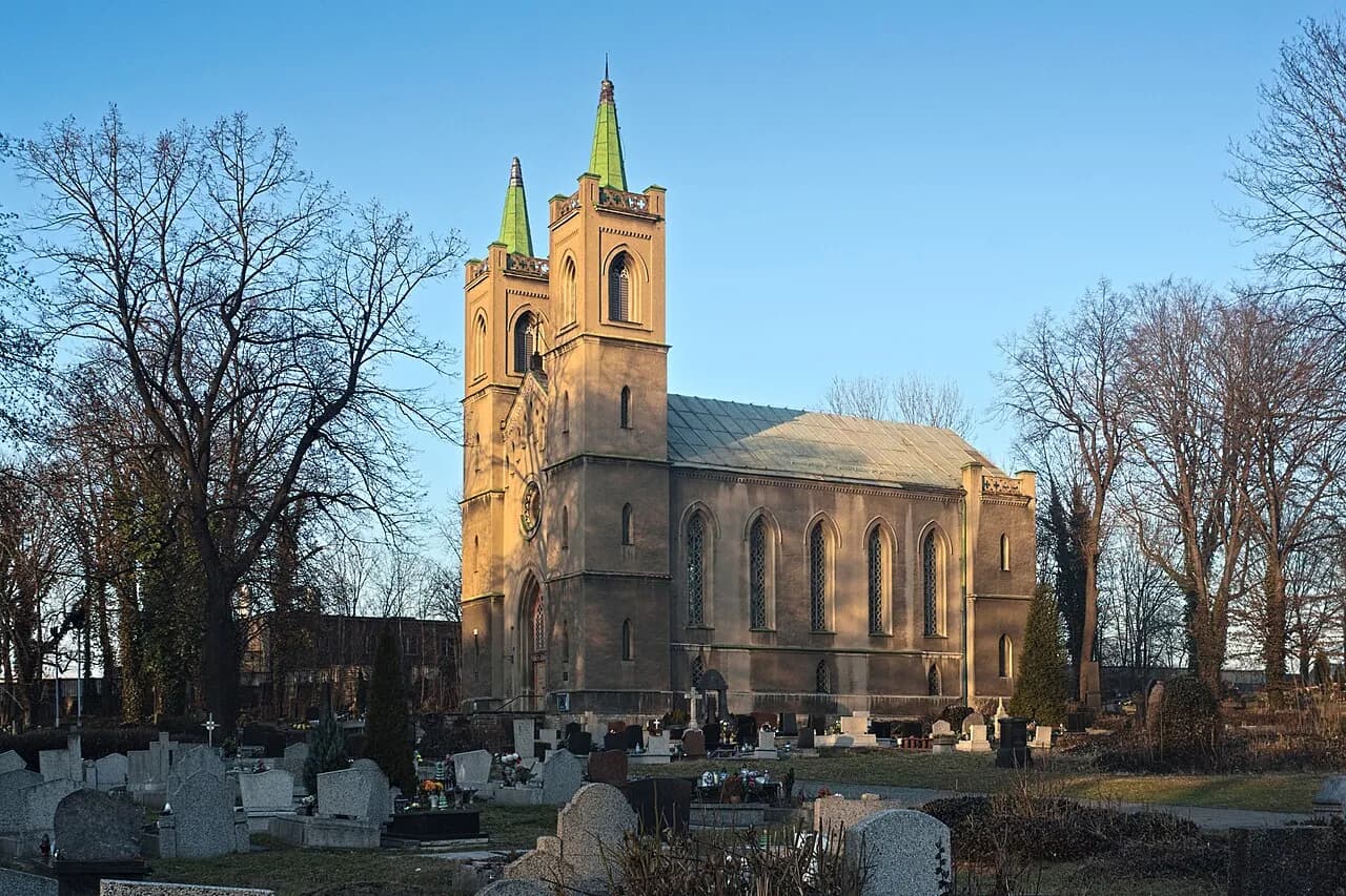 A large, old church with two prominent towers capped with green spires, stands in a cemetery under a bright blue sky, surrounded by bare trees in late afternoon sun.